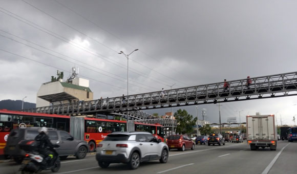 Красный трёхсекционный автобус из компании Трасмиленио ходит по выделенной линии, а к его остановке ведут пешеходные мосты через проезжую часть.